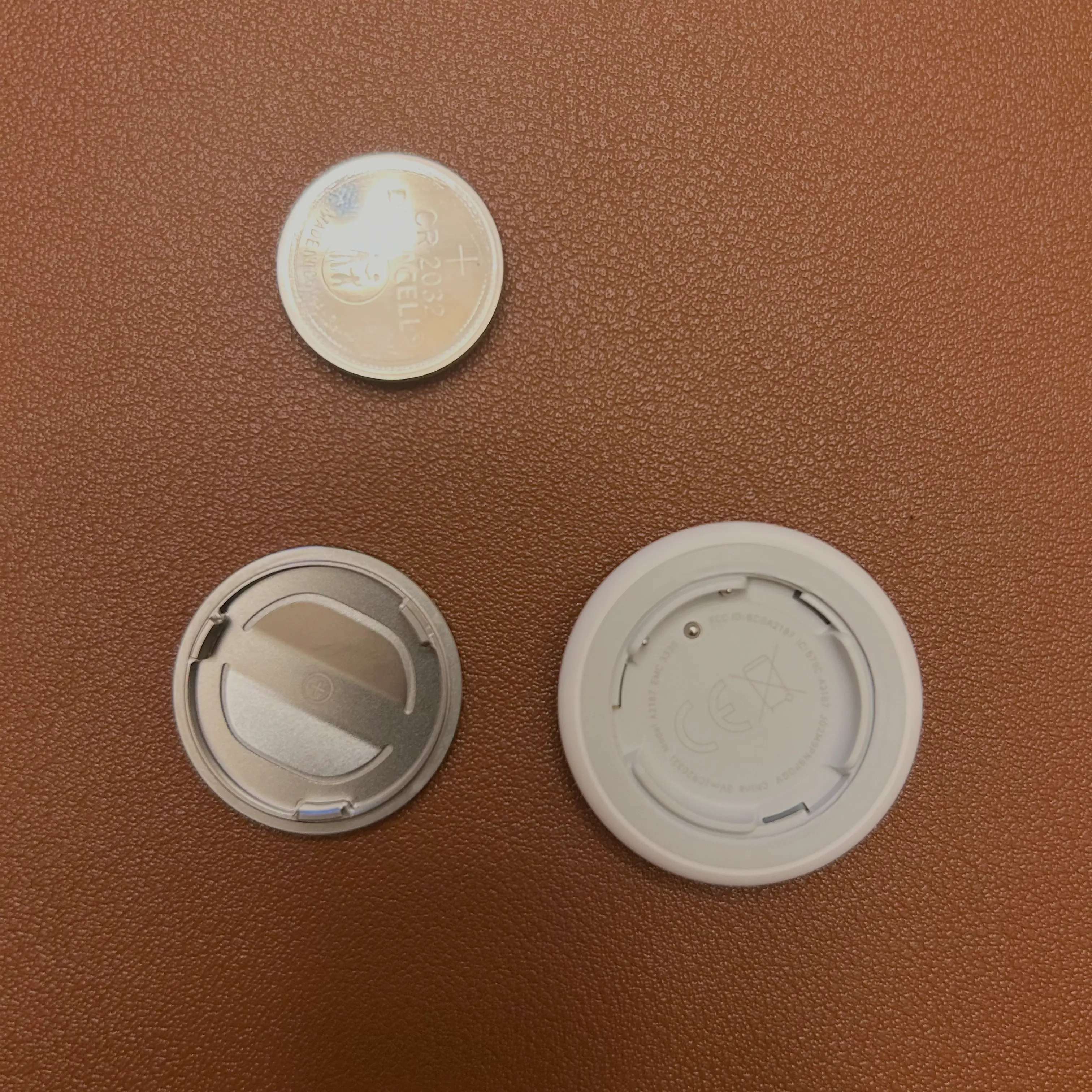 The AirTag's components laid out on a brown surface, including the metal back cover, the removed CR2032 battery, and the AirTag's plastic casing.